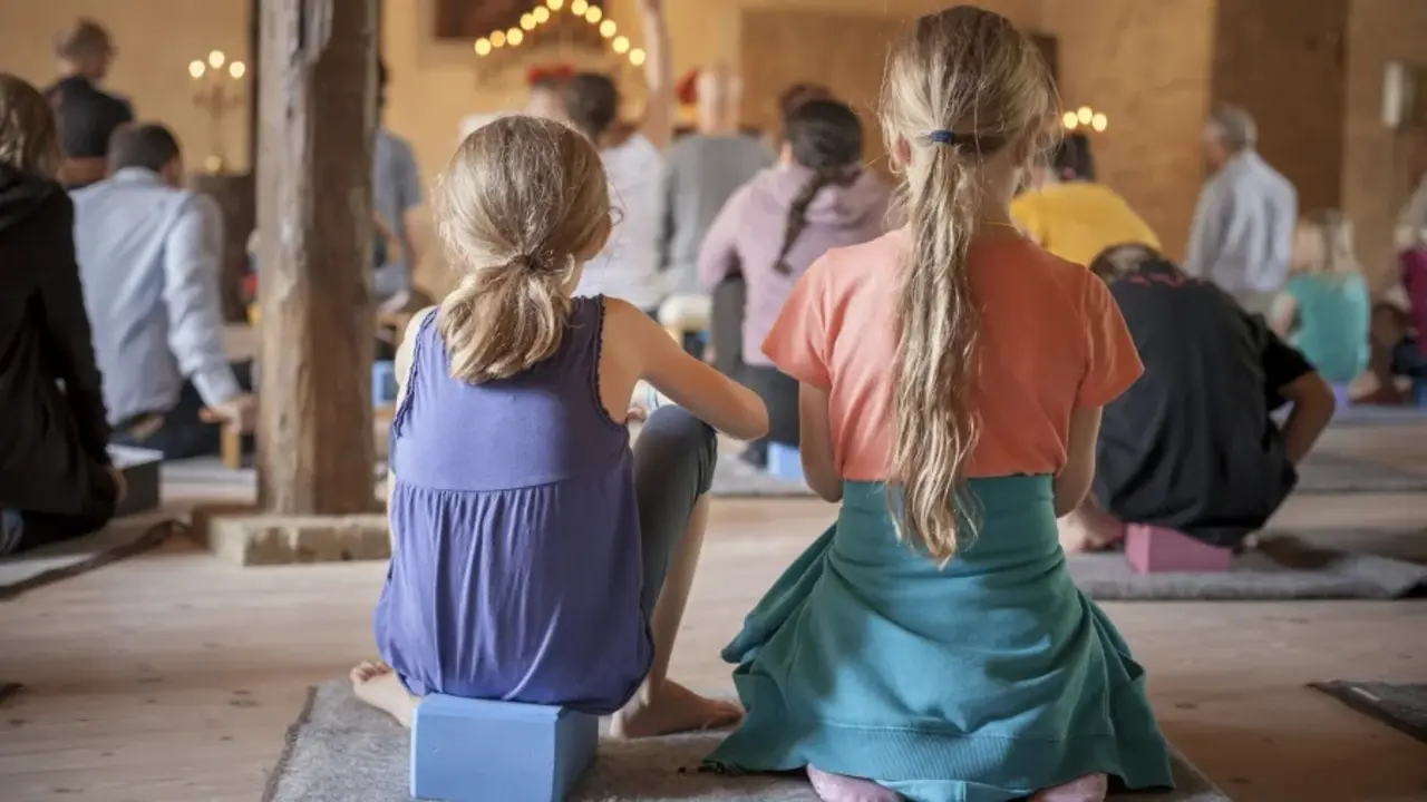 Rückenansicht von zwei Kindern, die in einem Meditationsraum mit Erwachsenen am Boden sitzen