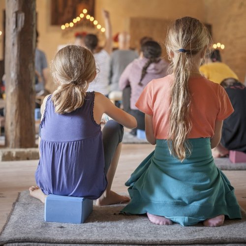 Rückenansicht von zwei Kindern, die in einem Meditationsraum mit Erwachsenen am Boden sitzen