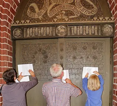 Vor dem Hauptportal der Lankwitzer Dreifaltigkeitskirche stehen drei Personen - zwei Erwachsene und ein Kind - und füllen einen Quizbogen zur Kirchenführung aus.